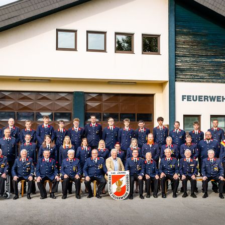 Eine Gruppe Feuerwehrleute in Uniform posiert für ein Foto vor einer Feuerwache. Das Schild sagt 'FEUERWEHR'. Sie tragen verschiedene Medaillen und Abzeichen auf ihren Uniformen.