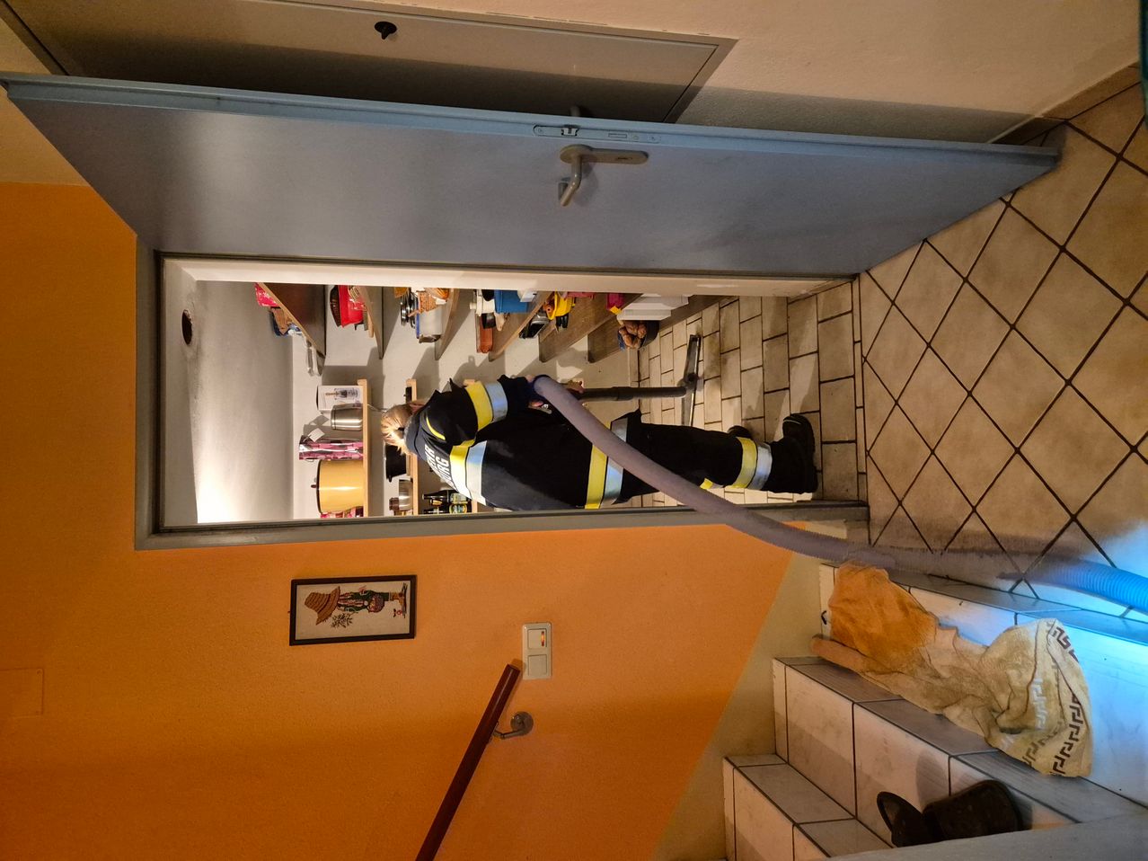 Ein Feuerwehrmann reinigt einen Bereich mit einem Staubsaugerschlauch und trägt dabei eine vollständige Ausrüstung. Der Raum hat geflieste Böden und Wände.