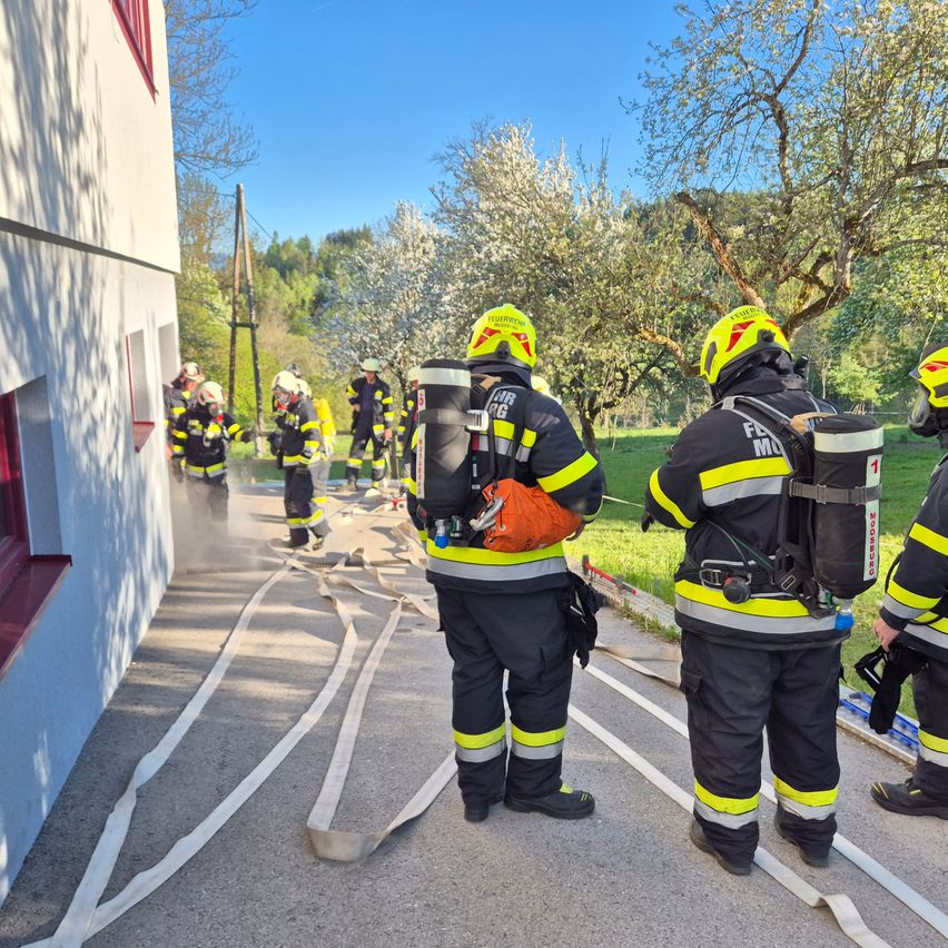 Eine Gruppe von Feuerwehrleuten in voller Ausrüstung übt an der Seite eines Gebäudes mit Feuerwehrschläuchen am Boden.