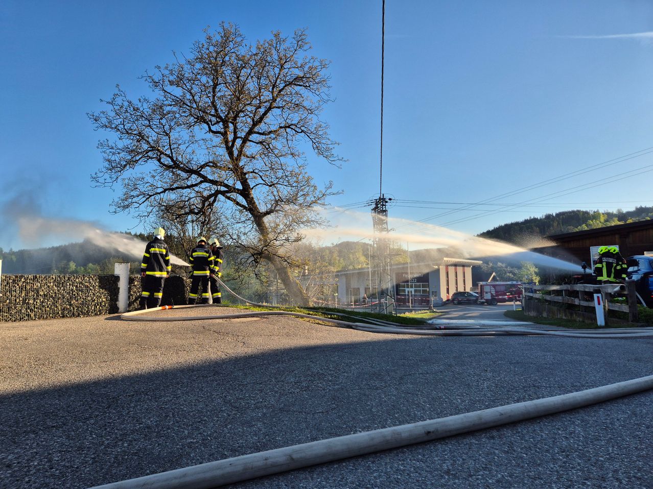 Feuerwehrleute stehen neben einer Straße und sprühen Wasser aus einem Schlauch. Ein Baum und ein Gebäude sind im Hintergrund.