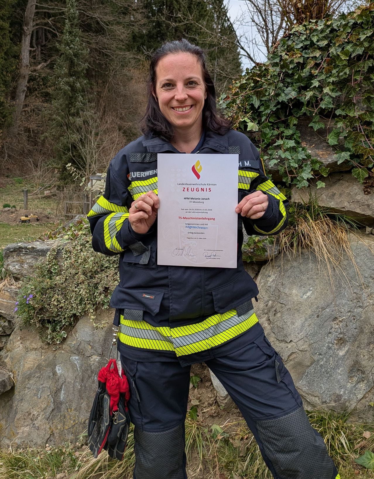 Eine Frau in einer Feuerwehruniform lächelt und hält ein Zertifikat. Das Zertifikat trägt den Aufdruck 'Zeugnis' und ist von 'Paul' unterzeichnet. Sie steht vor einer Steinmauer mit Pflanzen.