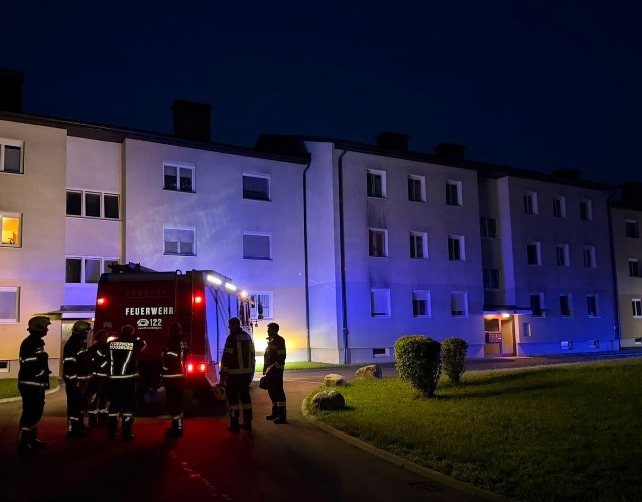Feuerwehrleute stehen nachts vor einem Gebäude, ein Feuerwehrwagen ist geparkt. Das Gebäude ist von blauen Lichtern beleuchtet.