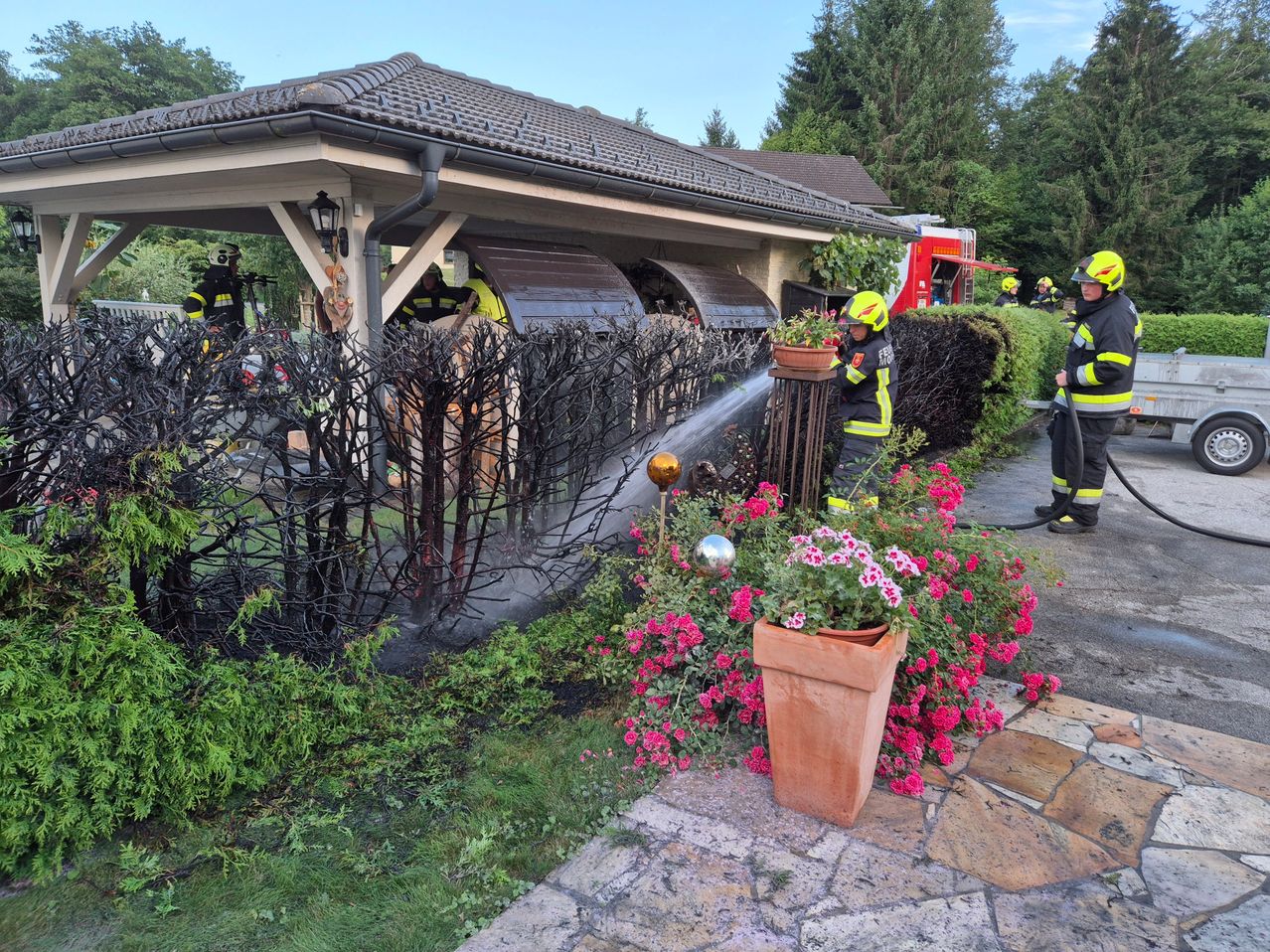 Feuerwehrleute löschen einen Brand in einem Gartenschuppen, einer benutzt eine Wasserspritze, während andere im Inneren arbeiten.