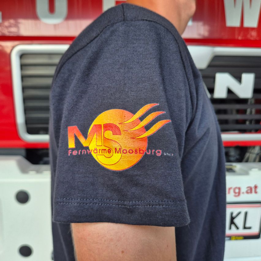 Der Rücken eines Menschen mit einem blauen Shirt, das ein Logo für Fernwärme Moosburg zeigt. Das Shirt hat eine Sonne mit Flammen und den Text 'Fernwärme Moosburg GmbH.' Das Bild wurde vor einem roten Feuerwehrauto aufgenommen.