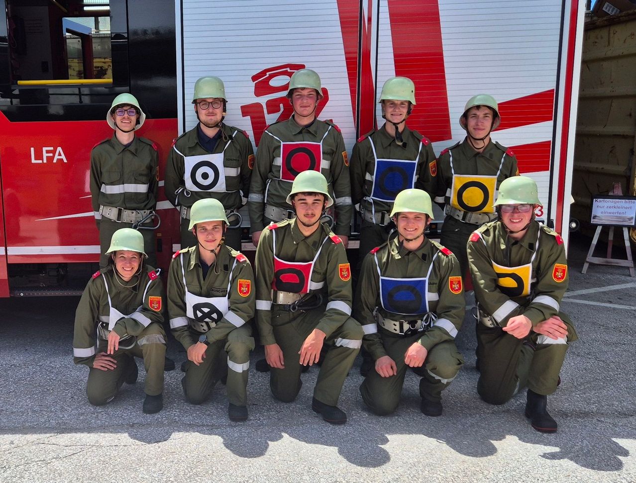 Eine Gruppe junger Menschen in Militäruniformen mit Helmen und Brillen posiert für ein Foto vor einem Feuerwehrwagen.