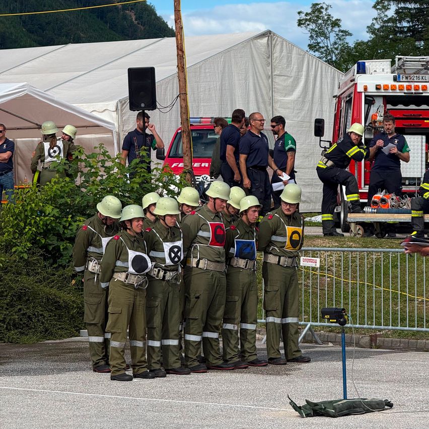Eine Gruppe Feuerwehrleute in Uniformen steht in einer Reihe mit Helmen und einem roten Kreuz auf der Brust. Dahinter unterhalten sich andere Leute in der Nähe eines Feuerwehrwagens. Zelte und ein Pfosten sind im Hintergrund zu sehen.