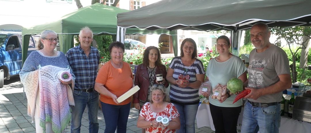 Eine Gruppe von Menschen steht unter einem Zelt, lächelt und hält Lebensmittel. Eine Frau hält eine Wassermelone und eine Frau neben ihr hält ein Glas. Eine Frau in der Mitte hält ein Kuscheltier. Ein Mann steht links.