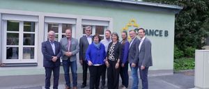 Eine Gruppe von Erwachsenen, möglicherweise Kollegen, steht vor einem Gebäude mit dem Wort 'ANCE' an der Wand. Sie alle lächeln und scheinen für ein Foto zu posieren.