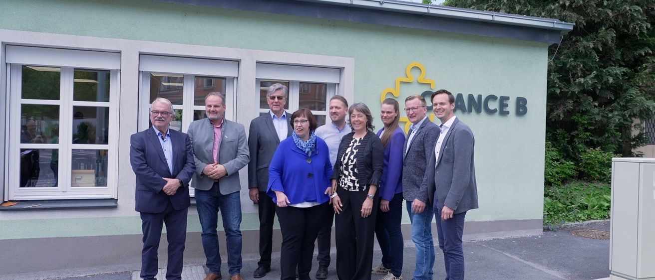 Eine Gruppe von sieben Personen, gekleidet in formeller Kleidung, steht vor einem Gebäude mit dem Schild 'SANCE'. Sie lächeln und scheinen für ein Foto zu posieren.
