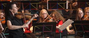 Eine Gruppe von Musikern spielt Geigen bei einem Konzert. Sie sitzen auf Stühlen, halten ihre Geigen und schauen auf ihre Notenblätter. Die Frau in der Mitte trägt eine Brille und hat ihre Haare zu einem Pferdeschwanz gebunden. Hinter ihnen spielen andere Musiker ihre Instrumente. Das Bild ist unscharf.