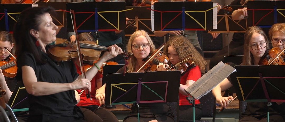 Eine Gruppe von Musikern spielt Geigen bei einem Konzert. Sie sitzen auf Stühlen, halten ihre Geigen und schauen auf ihre Notenblätter. Die Frau in der Mitte trägt eine Brille und hat ihre Haare zu einem Pferdeschwanz gebunden. Hinter ihnen spielen andere Musiker ihre Instrumente. Das Bild ist unscharf.