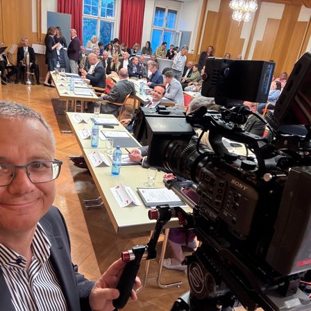 Ein Mann mit Brille hält eine Kamera in einem Konferenzraum, wo eine Sony-Kamera vor ihm aufgestellt ist. Hinter ihm ist ein Tisch mit Papieren und Flaschen zu sehen. Viele Menschen sitzen in Reihen, einige in formeller Kleidung, und andere stehen in der Nähe der Fenster.