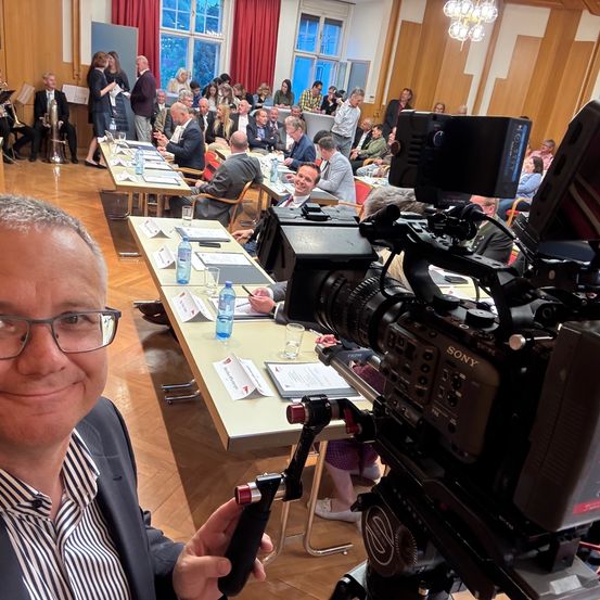 Ein Mann mit Brille hält eine Kamera in einem Konferenzraum, wo eine Sony-Kamera vor ihm aufgestellt ist. Hinter ihm ist ein Tisch mit Papieren und Flaschen zu sehen. Viele Menschen sitzen in Reihen, einige in formeller Kleidung, und andere stehen in der Nähe der Fenster.