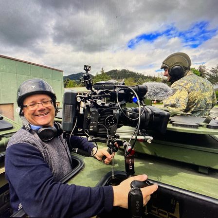 Zwei Männer in Militäruniformen mit Helmen filmen in einem grünen Fahrzeug mit einer Kamera und einem Mikrofon. Ein Mann lächelt und trägt eine Brille.