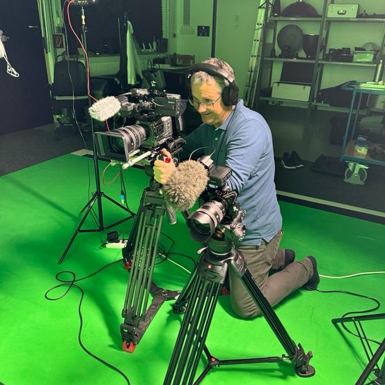Ein Mann in einem blauen Hemd kniet auf einem grünen Bildschirm in einem Studio und hält eine Kamera mit angebrachtem Mikrofon.