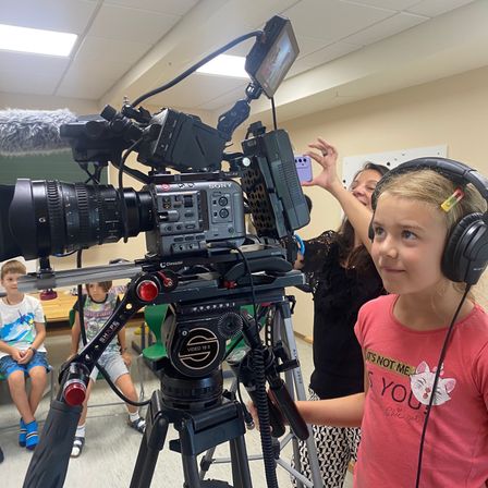 Ein junges Mädchen mit Kopfhörern beobachtet eine Videokamera auf einem Stativ, während andere Kinder im Hintergrund sitzen.