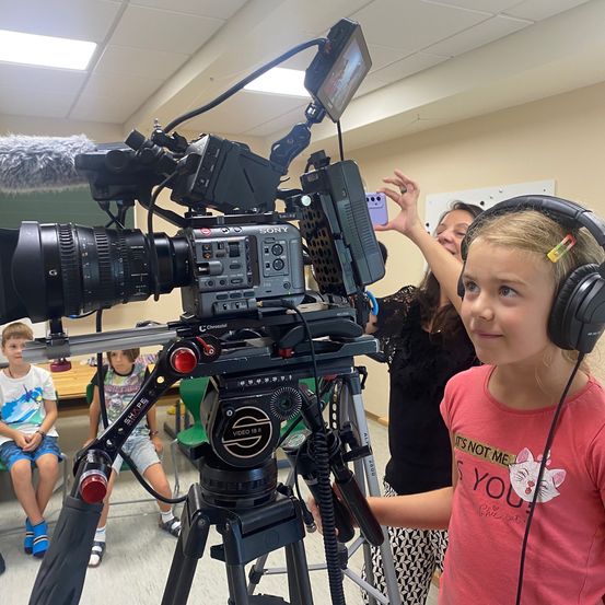Ein junges Mädchen mit Kopfhörern beobachtet eine Videokamera auf einem Stativ, während andere Kinder im Hintergrund sitzen.