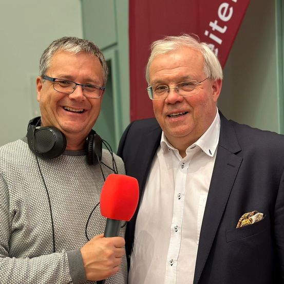Zwei Männer lächeln, einer hält ein Mikrofon. Beide sind formell gekleidet, der linke trägt eine Brille und Kopfhörer, der rechte einen Anzug mit Brusttasche. Dahinter befindet sich ein roter Banner mit weißem Text.