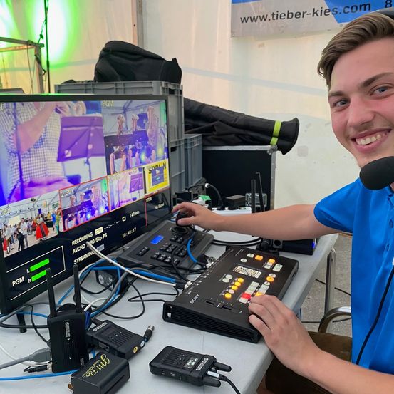 Ein junger Mann bedient eine Videomischkonsole, lächelt und ist von Monitoren und Geräten umgeben. Die Umgebung scheint ein Studio zu sein mit verschiedenen Kabeln und Geräten auf dem Schreibtisch.