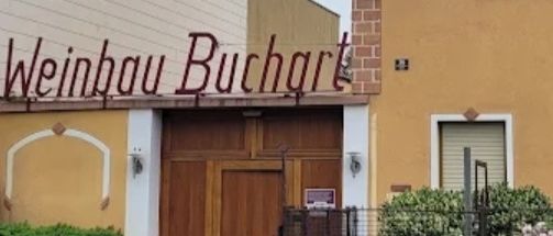Ein gelbes Gebäude mit einer hölzernen Tür, beschriftet mit 'Weinbau Buchart', steht neben einer Straße mit Pflanzen und einem Zaun.