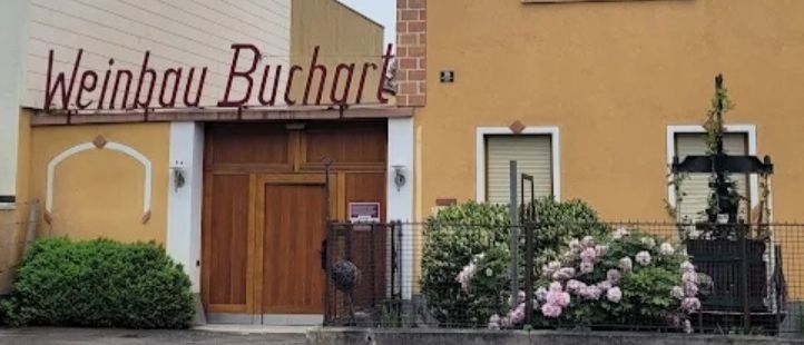 Ein gelbes Gebäude mit einem hölzernen Eingang, der mit Weinhau Buchart beschriftet ist. Es hat rote Buchstaben und einen kleinen Garten mit Blumen.