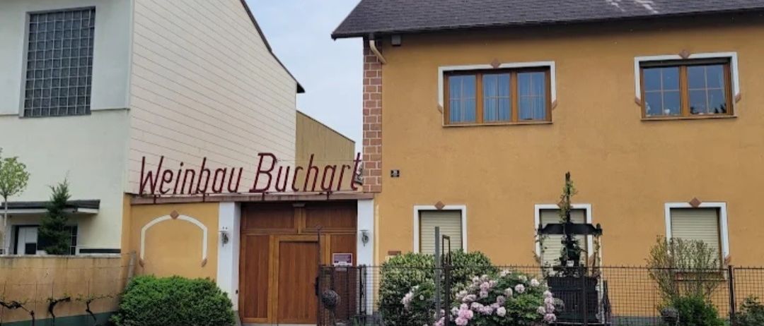 Ein gelbes Haus mit einem hölzernen Eingang, Weinhau Buchart darüber und einem Garten mit rosa Blumen und Sträuchern davor.