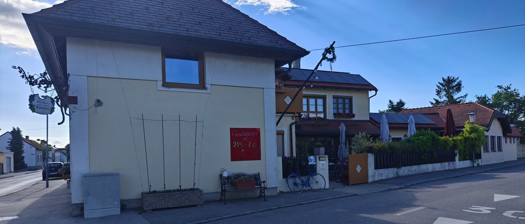 Ein Haus mit einem roten Schild 'Pension' und einer Bank davor, ein Fahrrad ist in der Nähe geparkt. Bäume und Pflanzen stehen vor dem Haus. Der Himmel ist blau mit Wolken.