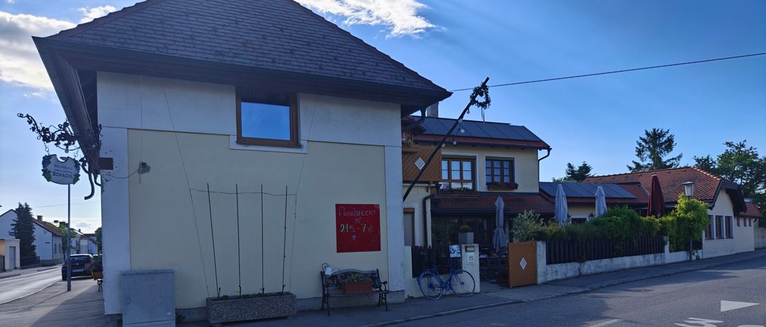 Ein Haus mit einer Bank und einem Fahrrad davor. Das Gebäude hat ein rotes Schild mit der Nummer 215 und Text. Es gibt Pflanzen und einen Zaun. Der Himmel ist klar.