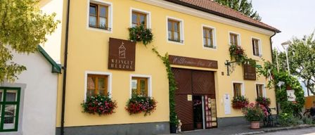 Ein gelbes Gebäude mit braunen Akzenten und mehreren Fenstern. Es hat eine braune Tür mit einem Schild, das Weingut Herzog besagt. Vor den Fenstern befinden sich Blumenarrangements und ein Topfpflanzen neben dem Eingang.