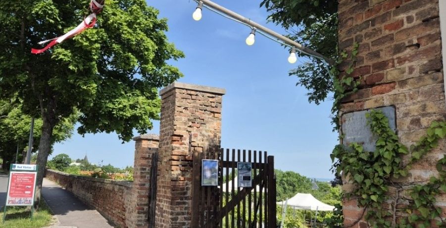 Ein Torbogen aus Ziegelsteinen mit einem dekorativen Tor, beleuchtet von Lampen, führt in einen Garten. Ein Weg verläuft neben einer Ziegelsteinmauer, und ein weißes Zelt ist in der Ferne zu sehen.