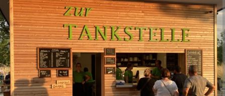 Eine Gruppe von Menschen steht vor einem kleinen Laden mit dem Schild 'zur Tankstelle'. Ein Menü ist an der Wand und ein Schild 'Leergut DANKE' ist an der Wand.