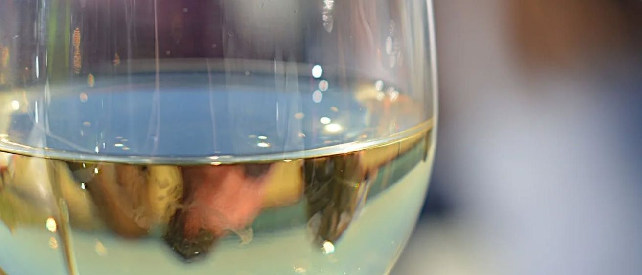 Ein Nahaufnahme eines Weinglases, gefüllt mit klarem Weißwein. Das Glas ist halbvoll, und die Reflexion einer verschwommenen Person und der Umgebung sind auf dem Glas zu sehen.