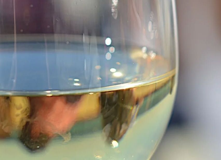 Ein Nahaufnahme eines Weinglases, gefüllt mit klarem Weißwein. Das Glas ist halbvoll, und die Reflexion einer verschwommenen Person und der Umgebung sind auf dem Glas zu sehen.