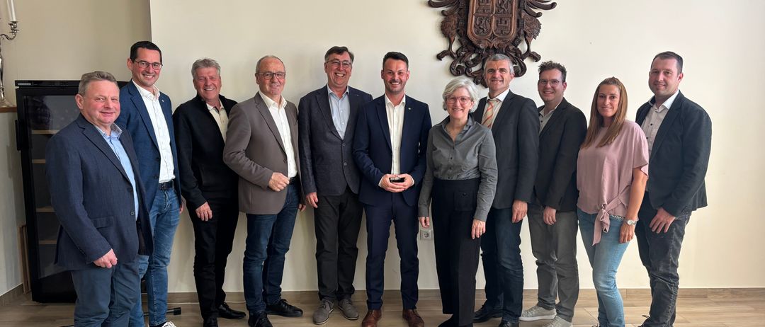 Eine Gruppe von Menschen in Geschäftskleidung steht in einem Raum und lächelt für ein Foto vor einer weißen Wand mit einem dekorativen Emblem.