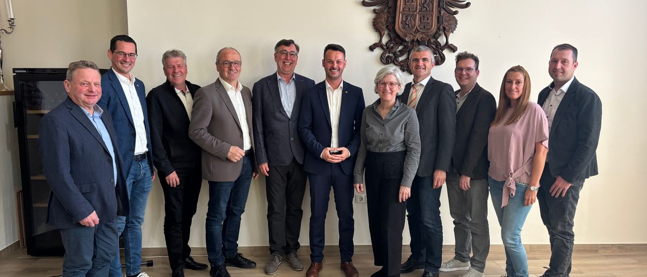 Eine Gruppe von Menschen in Geschäftskleidung steht in einem Raum und lächelt für ein Foto vor einer weißen Wand mit einem dekorativen Emblem.