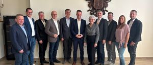 Eine Gruppe von Menschen in Geschäftskleidung steht in einem Raum und lächelt für ein Foto vor einer weißen Wand mit einem dekorativen Emblem.