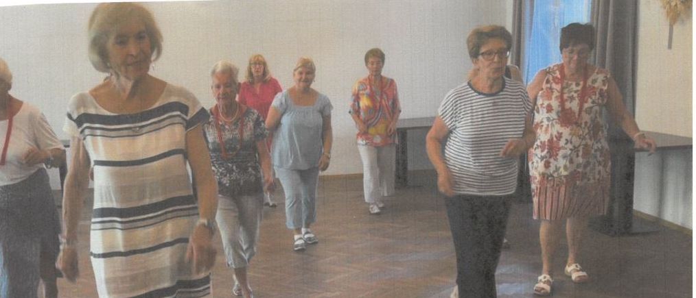 Eine Gruppe älterer Frauen tanzt in einem Raum. Die Frau vorne trägt ein gestreiftes Kleid. Der Raum hat einen Holzboden und eine weiße Wand. Es gibt zwei Tische an der Seite. Der Text oben ist auf Deutsch.