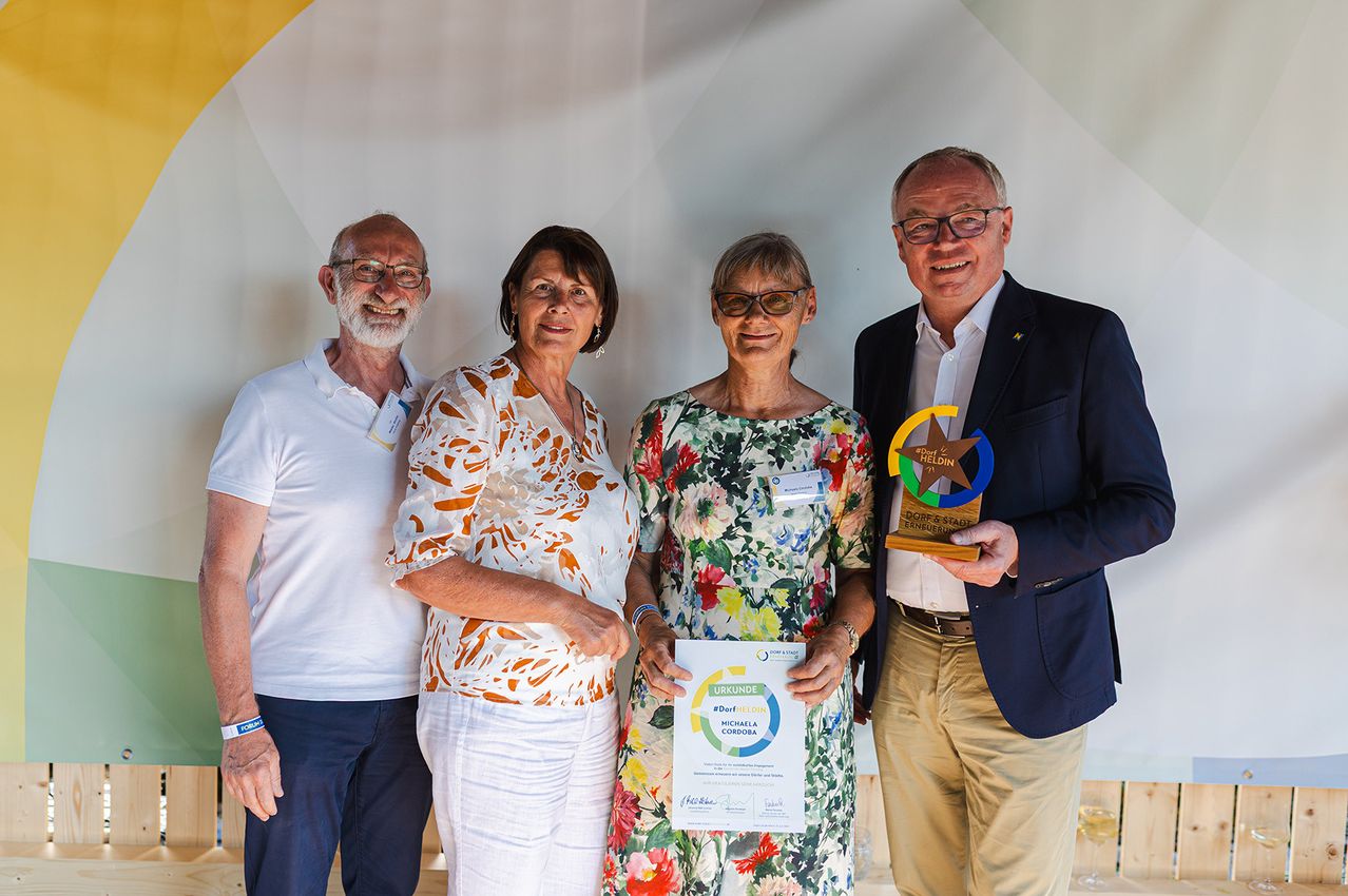 Vier Erwachsene, zwei Männer und zwei Frauen, posieren für ein Foto mit Auszeichnungen. Die Frau in der Mitte hält ein Zertifikat. Der Mann rechts hält einen Pokal.