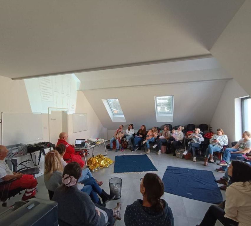 Ein Innenraum mit hoher Decke, einer Projektionswand und mehreren Personen, die auf Stühlen sitzen. Einige Menschen halten Säuglinge, während andere verschiedene Aktivitäten durchführen. Der Raum ist gut beleuchtet, mit natürlichem Licht, das durch Fenster einfällt.