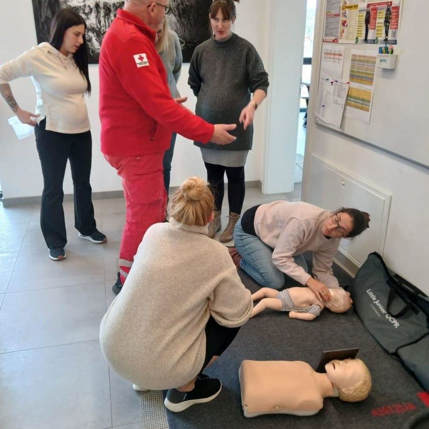 Eine Gruppe von Menschen in einem Raum übt CPR an einem Dummy. Eine Frau kniet und führt Brustkompressionen durch, während eine andere Frau steht und zusieht.