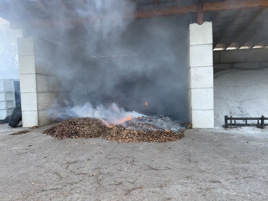 Ein Feuer brennt Blätter und Abfälle in einem Schuppen und gibt Rauch ab. Der Schuppen hat ein Holzdach und Betonwände.