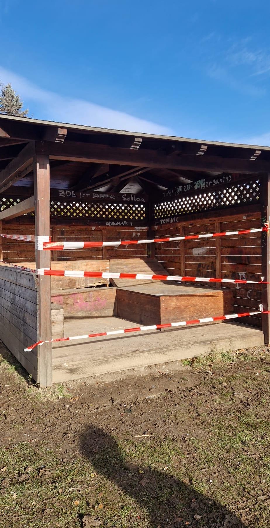 Eine Holzstruktur mit Dach und Wänden, möglicherweise ein Schuppen, mit Graffiti an den Wänden. Ein Holzstuhl ist drinnen und ist mit rot-weißem Klebeband abgesperrt.