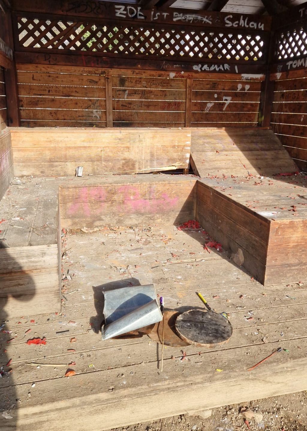 Eine alte Holzstruktur, möglicherweise ein Schuppen, mit Graffiti an den Wänden. Holzbretter sind auf dem Boden verstreut mit Überresten von roter Farbe. Ein hölzerner Hocker und eine gebrochene Holzplanke sind ebenfalls vorhanden.