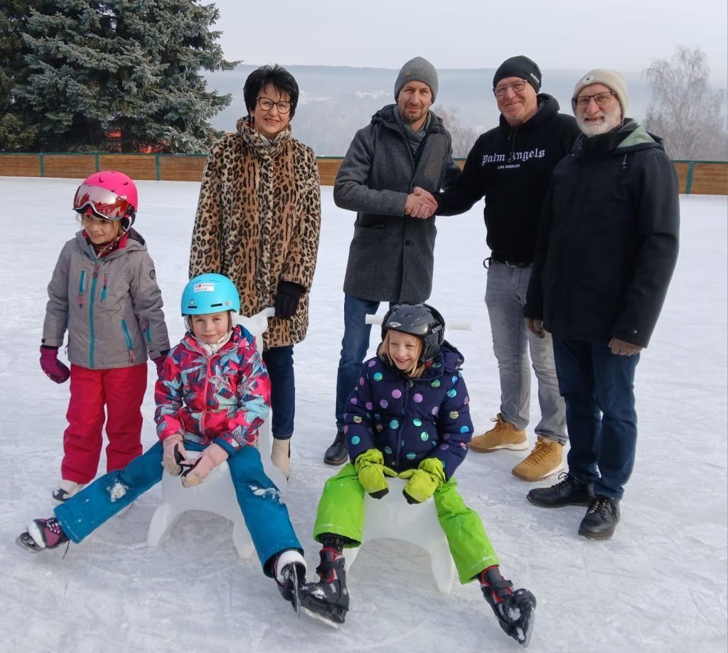 Eine Familie aus fünf Erwachsenen und drei Kindern in Winterkleidung steht auf einer Eisbahn, wobei zwei Erwachsene stehen und die anderen auf weißen Stühlen sitzen.