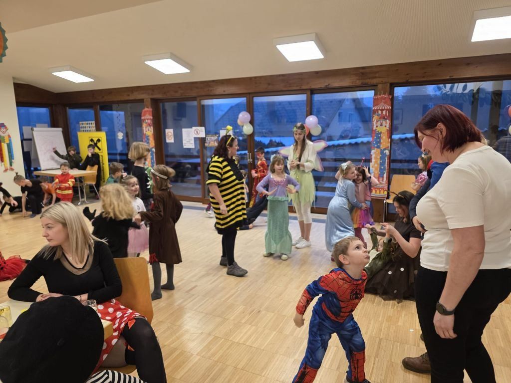Eine Gruppe von Kindern in Kostümen tanzt in einem Raum mit Erwachsenen. Ein Kind trägt ein Spiderman-Kostüm. Der Raum hat große Fenster und Luftballons.