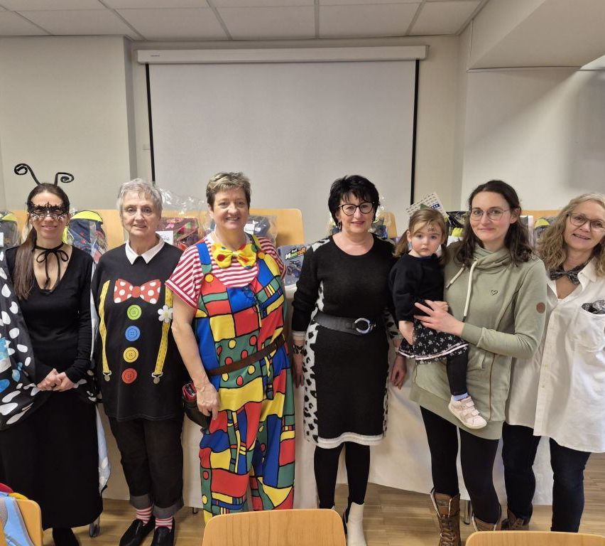 Eine Gruppe von Frauen und einem Kind in Clown-Kostümen posiert für ein Foto in einem Raum. Dahinter steht ein Tisch mit verschiedenen Gegenständen und eine Projektionsleinwand an der Wand.