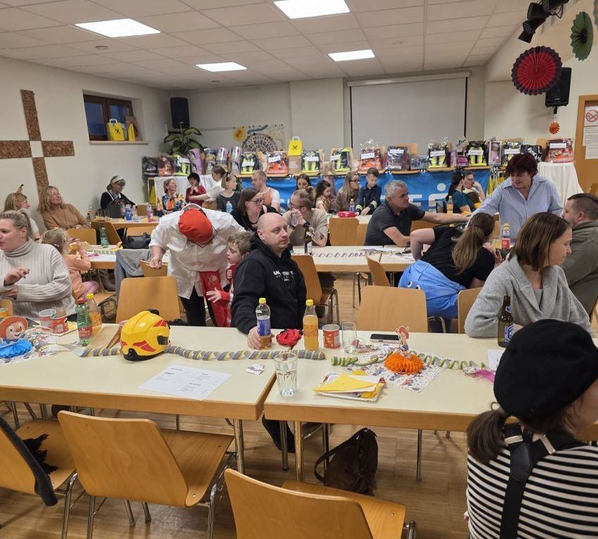 Eine Gruppe von Menschen ist in einem Raum für eine Party versammelt. Tische sind mit Flaschen, Tellern und Gläsern gedeckt. Ein Mann trägt einen roten Hut.