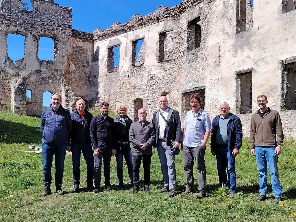 Eine Gruppe von Menschen steht vor einem zerstörten Gebäude an einem sonnigen Tag. Sie alle lächeln und scheinen für ein Foto zu posieren.