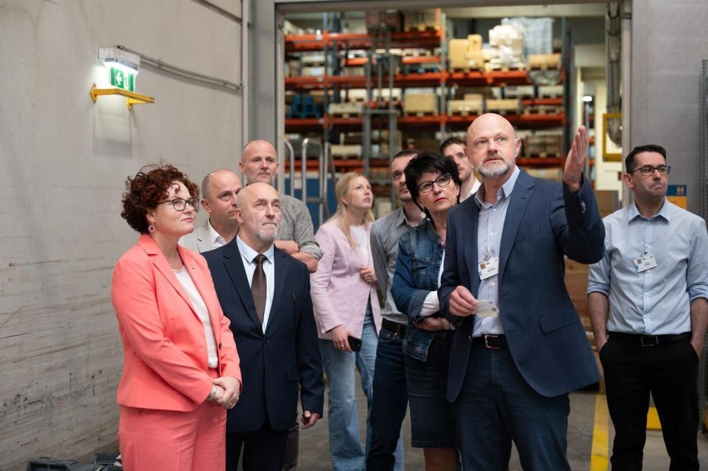 A group of formally dressed adults stand in a warehouse, listening to a man giving a presentation.