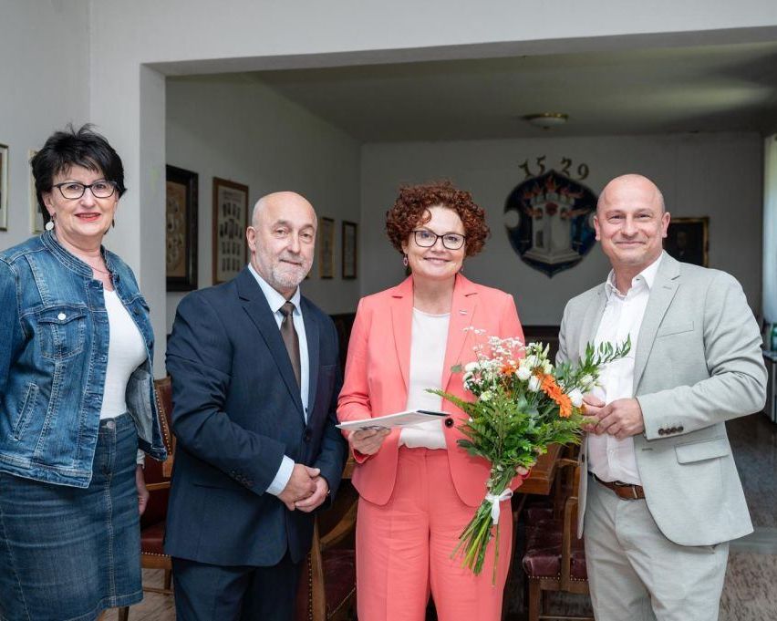 Four people stand together in a room, smiling for a photo. A woman holds flowers and a document. Behind them is a wall with a logo.